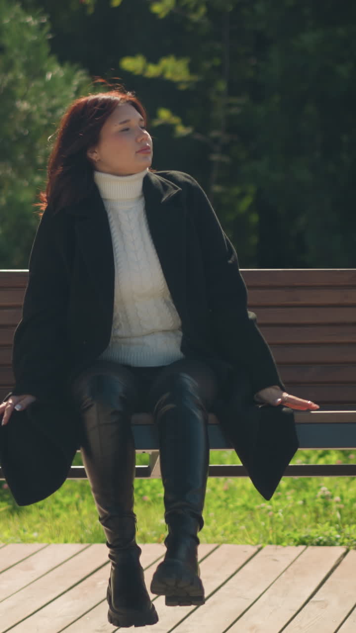 젊은 여성은 은 공원에서 목조 스에 앉아 머리를 조정합니다, 세련된 검은 코트와 부츠를 입고, 울창한 초록색으로 둘러싸여, 조용한 표정과 손을 스에 기대고 있습니다.