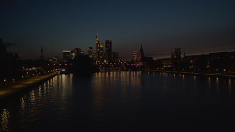 wide view of frankfurt city skyline at dusk. skyscrapers and historic buildings are lit up, reflecting on the river