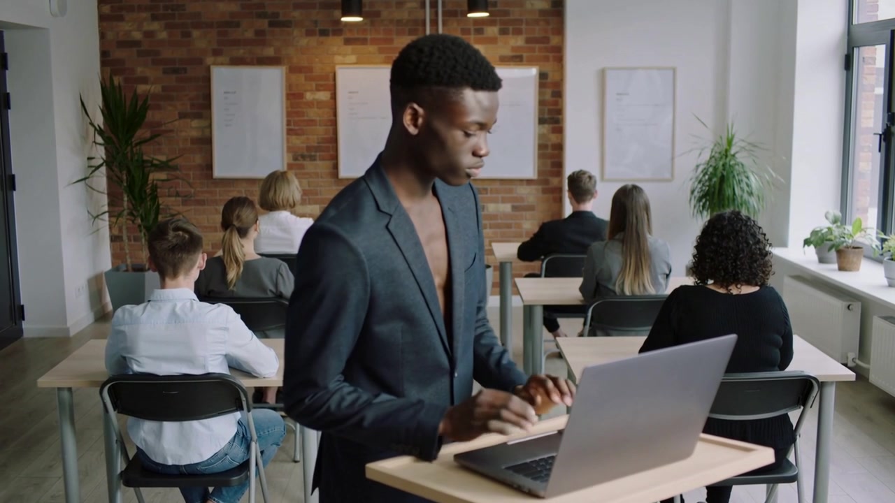 Talende person i mørk blazer taster, kigger til venstre, vender sig, går frem til klassen, bærbar computer på talerstol.