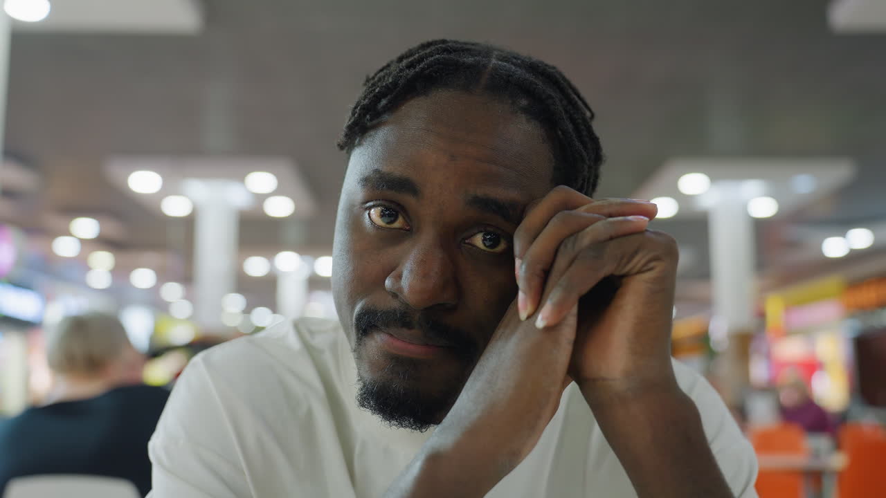 Close up of calm african man resting head on folded hands with eyes closed sitting quietly in crowded food court surrounded by blurred lights showing peace meditation