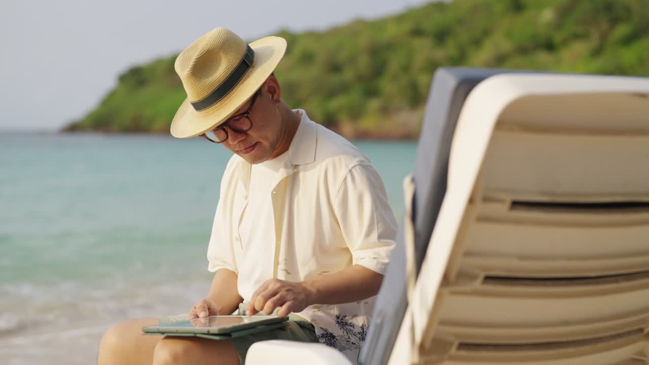 empresario asiático feliz trabajando en una tableta digital mientras viaja en una playa tropical.