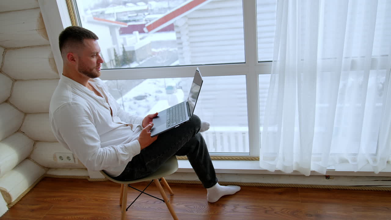Focused bearded man works on his laptop sitting at the window. Person working remotely away from office.