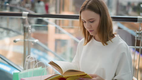 joven sentada en un centro comercial moderno leyendo un libro amarillo con enfoque y reflexión bolsas de compras a su lado reflejan una pausa de compras relajada en un ambiente de centro comercial bien iluminado con suave elegante