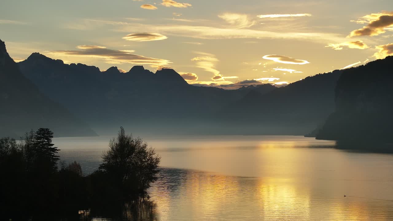 Misty sunrise over Walensee, with golden light reflecting on calm waters and mountains silhouetted against the sky in Switzerland