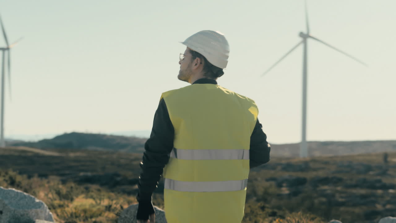 Walking among wind turbines, a young Caucasian engineer in a white helmet and vest represents the growth of clean energy for a greener future