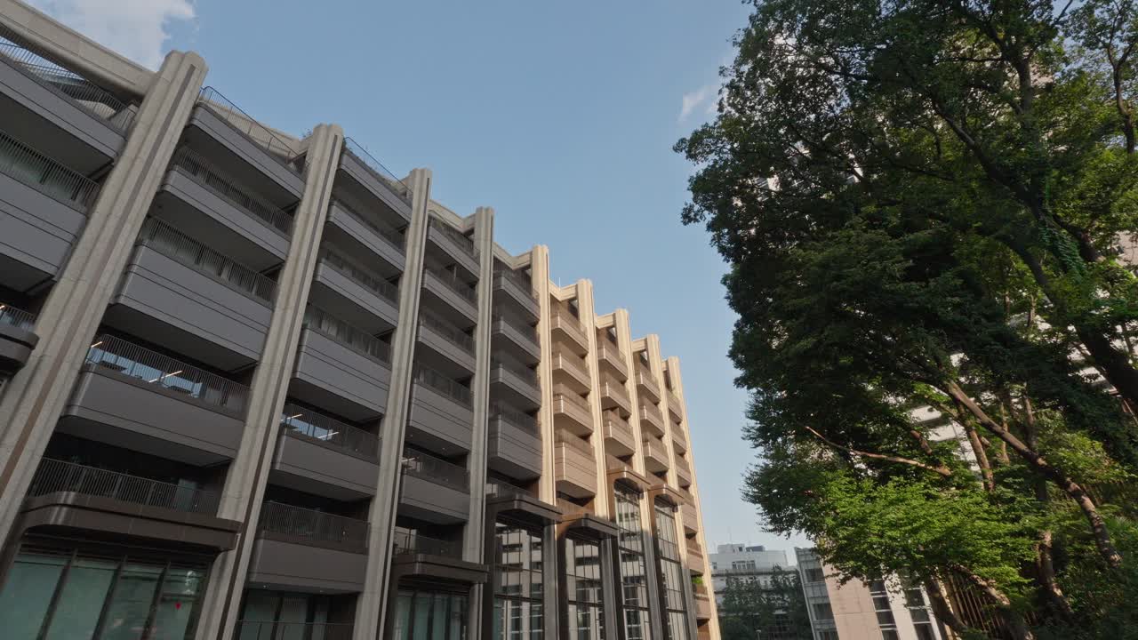 A modern building with stacked balconies, likely in the Azabudai Area, stands beside tall, leafy trees under a clear blue sky.