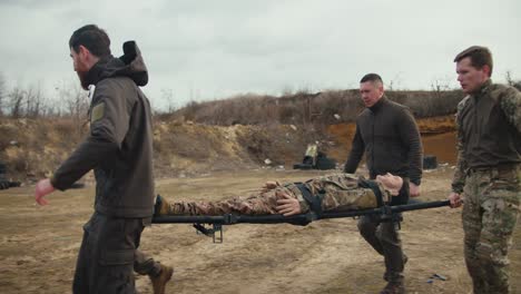 Side-view-of-four-confident-male-soldiers-in-dark-green-uniforms-carrying-a-wounded-doll-of-a-male-soldier-in-camouflage-uniform-on-a-stretcher-and-placing-him-on-the-ground-near-an-ambulance-on-a-training-ground-during-a-military-medicine-exercise