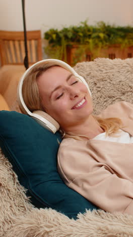 woman lying on sofa with eyes closed listening to music through headphones in peaceful relaxation
