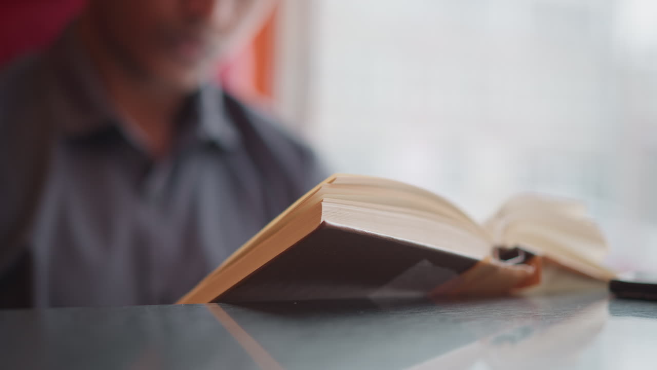 Young man reading hardcover book indoors with soft daylight coming through window, focused on pages while sitting calmly, wearing button-up shirt, engaged in quiet learning or study moment