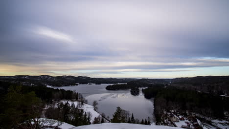 Time-lapse-shot-of-dark-clouds-above-a-lake,-hills-and-forest,-on-a-sunny,-winter-sunset,-in-Grimenes,-Agder,-South-Norway