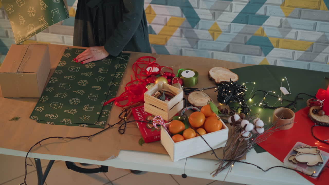 Cozy christmas present wrapping on table, hands arrange green patterned paper, kraft box, red ribbon and fairy lights, wooden crate with oranges and cotton sprigs, warm ambient lighting, artisan gift