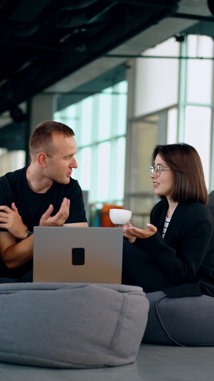 Active communication between the colleagues in the office. Man and woman sit in bean bag chairs in front of laptop having discussion. Vertical video.