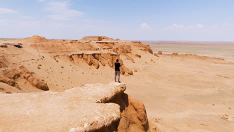 Aerial-Revealed-Man-Standing-Over-Flaming-Cliffs-Bayanzag-In-Gobi-Desert,-Mongolia