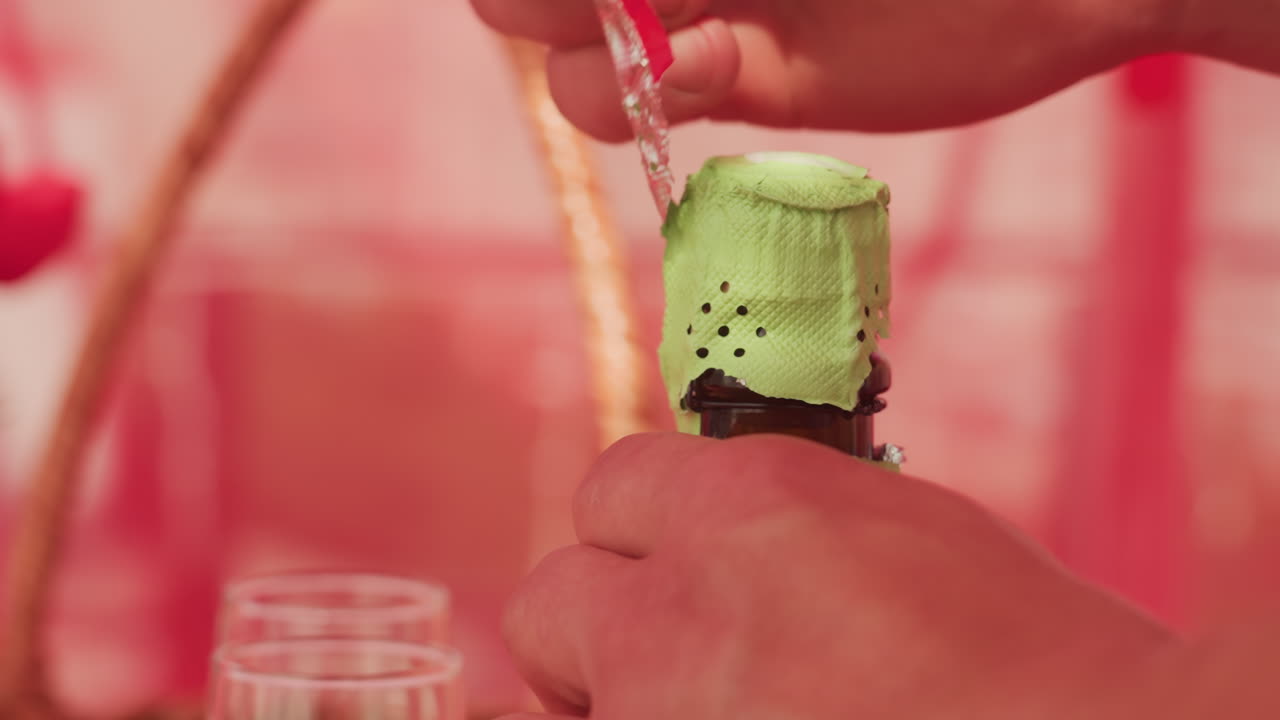 Mani che stappano champagne a un picnic, primo piano sulla stagnola verde e sulla gabbia metallica mentre mani attente rimuovono la stagnola e sollevano la gabbietta; il tappo è pronto a saltare, due calici e un cesto di vimini sono sfocati nella calda luce del sole