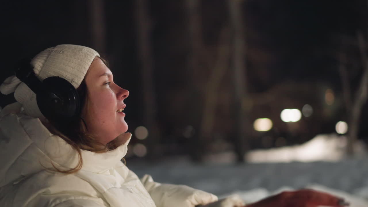 Female singer seated in snowy park at night, wearing white winter coat and headphones, eyes closed in peaceful moment, embracing winter atmosphere and enjoying music in bright outdoor lighting