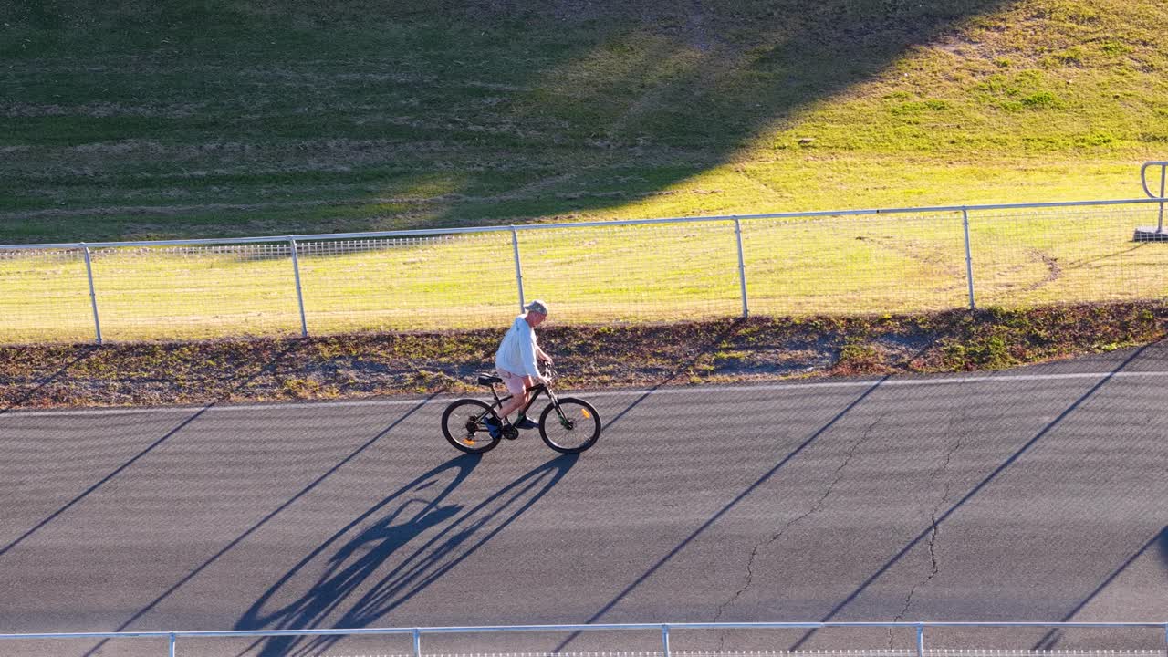 A teenager rides a bicycle along a sunlit track, casting long shadows on the pavement in a park setting