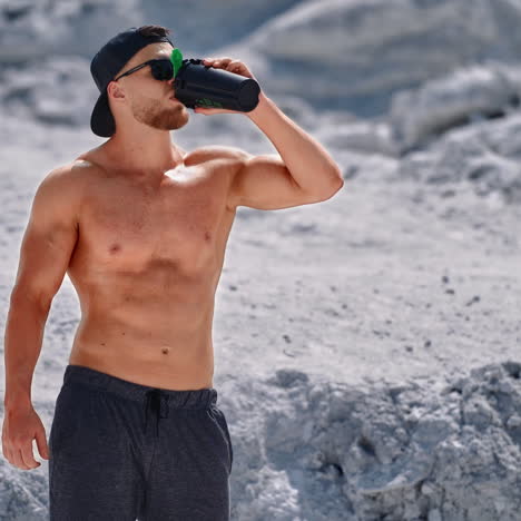 topless sportsman drinking cold drink on natural background. muscular man in black sunglasses drinks water after workout in the mountains.