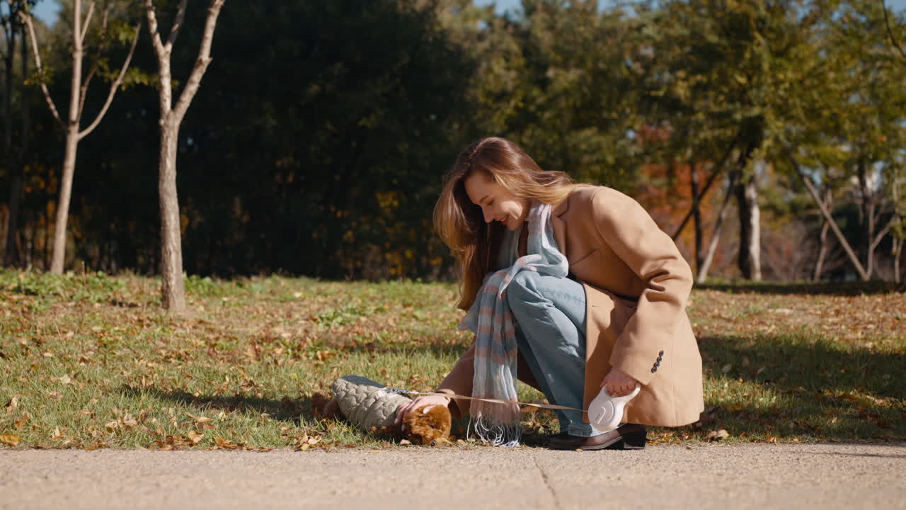 Happy woman is kneeling on the ground, smiling as she tickles a small Toy poodle dog on a leash during a walk in Autumn Park in slow motion