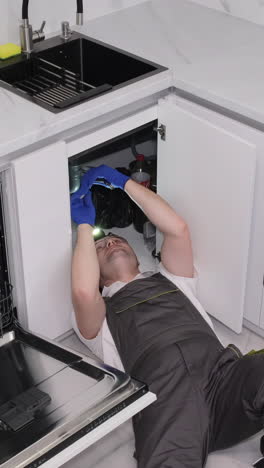 a male plumber in overalls is repairing a water leak from a sink in the kitchen the work of a
