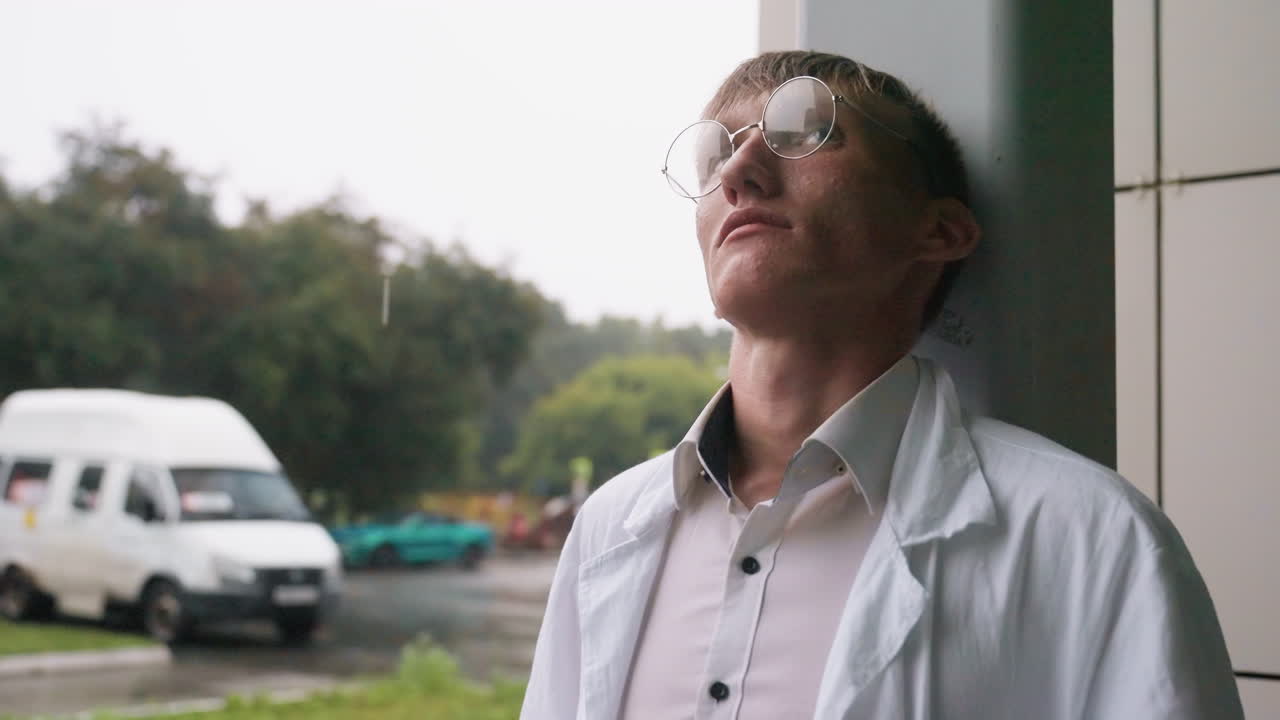 Young biologist in white coat leans head against wall during outdoor break, eating pastry with thoughtful expression in calm urban setting with blurred view of cars and trees in background