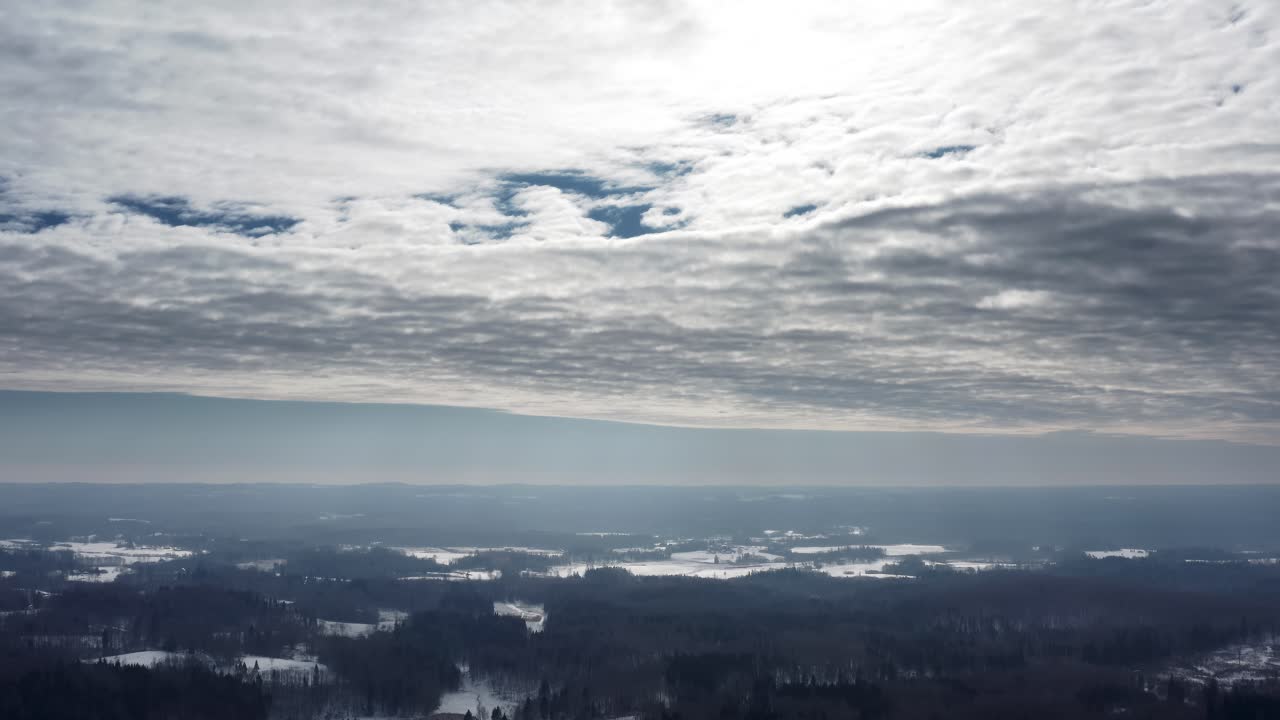 Aerial drone view high up in the sky over a snow covered countryside landscape in winter. Sunrays through clouds over a dense forest.