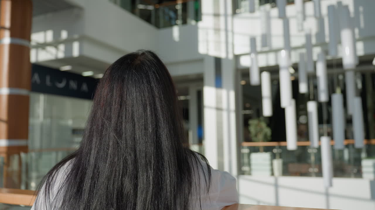 Back view of woman with long black hair walking toward balcony rail inside spacious modern mall with stylish cylindrical light fixtures and glass panels in bright natural light