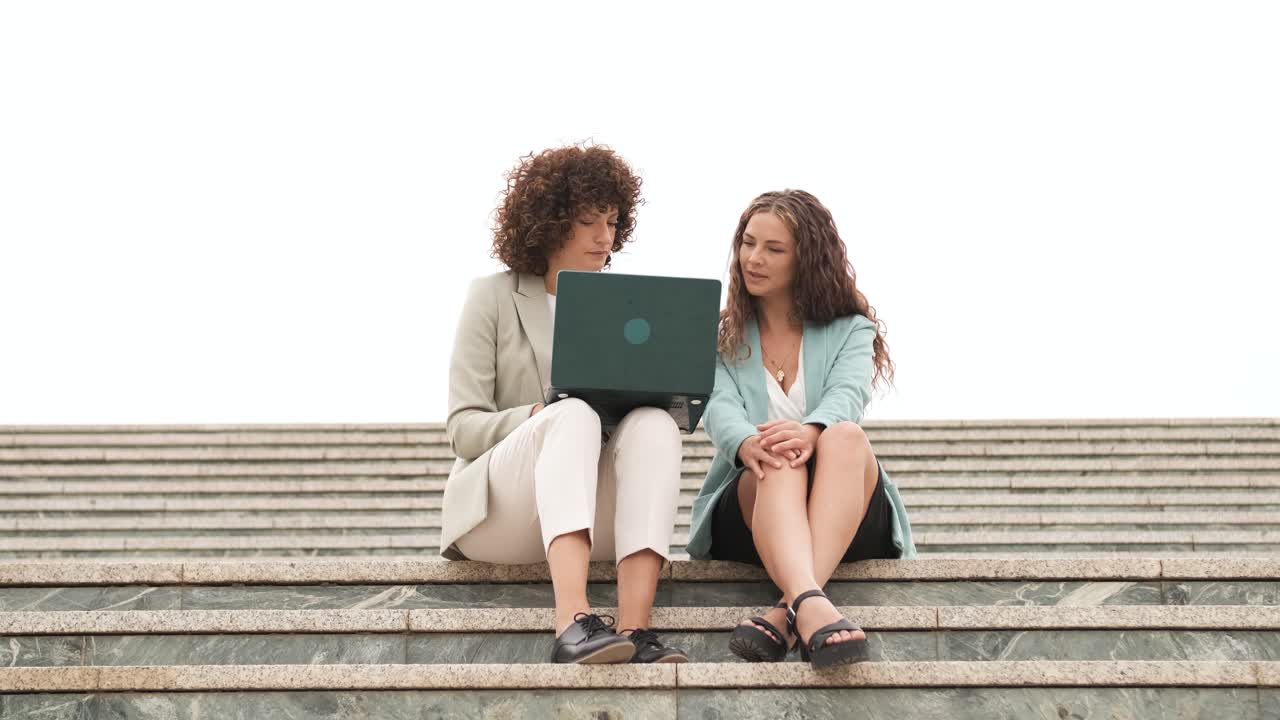 Smiling businesswomen using netbook and talking on steps in city