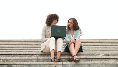 smiling businesswomen using netbook and talking on steps in city