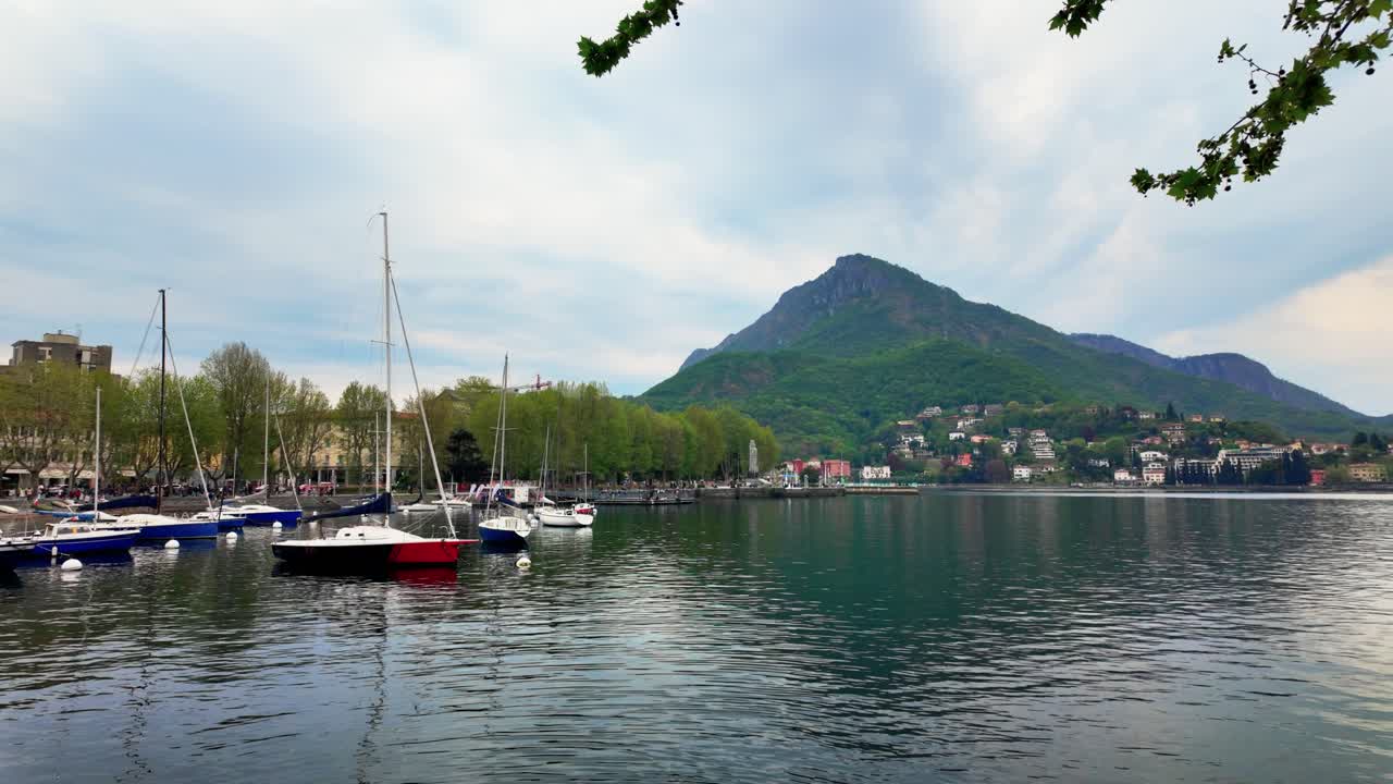Riprese grandangolari del pittoresco lungolago di Lecco sul Lago di Como con barche a vela ormeggiate, alberi verdi lussureggianti, riflessi tranquilli, montagne distanti e cieli diurni nuvolosi.