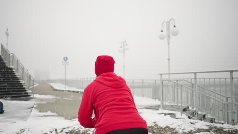 dama realizando jump squats durante el invierno entrenamiento al aire libre en terreno nevado usando sudadera roja con capucha y guantes, el fondo incluye bolsa en el suelo, barandilla de hierro, escalera, colina nevada y atmósfera brumosa