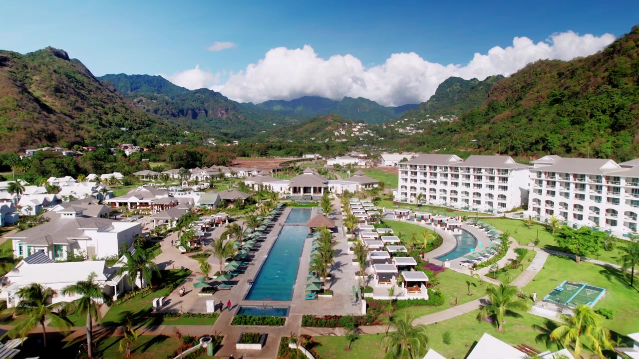 Ripresa aerea con drone a bassa quota che si sposta verso l'interno dal Mar dei Caraibi, sorvolando la spiaggia e l'area della piscina, rivelando l'architettura del resort di lusso, i giardini paesaggistici e il territorio tropicale montuoso.