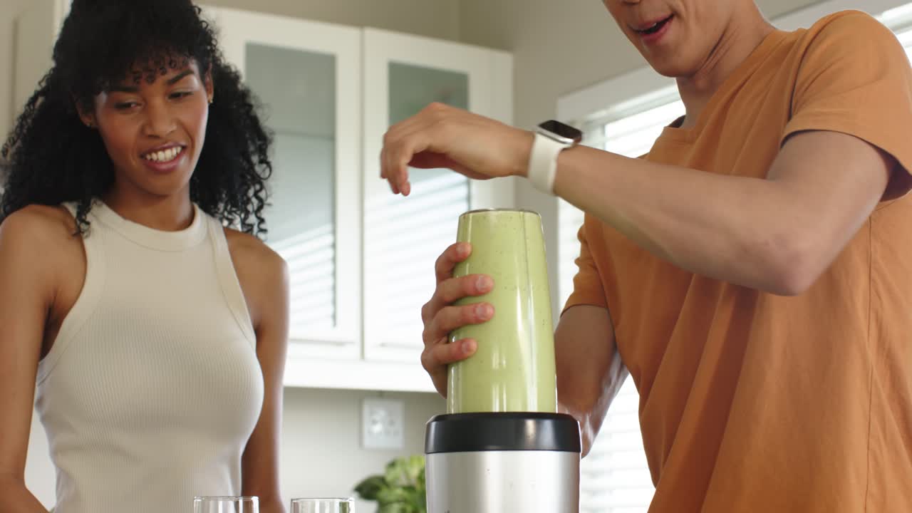 Mangfoldig par på kjøkkenet som trykker på smartklokke, fester glasset, blander grønn smoothie og heller opp drikke