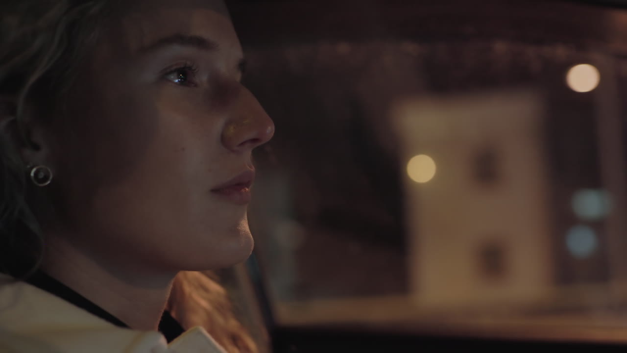 Close up of woman seated inside moving car at night, softly illuminated by city lights, with blurred view of building structure visible outside window, during late night drive