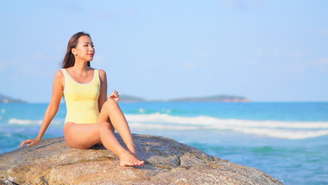 Schöne-Asiatische-Frau-In-Gelbem-Monokini,-Die-Auf-Einem-Großen-Felsen-Am-Meer-Auf-Der-Karibikinsel-Sitzt-Und-Ihr-Haar-Berührt,-Slow-Motion-Handheld