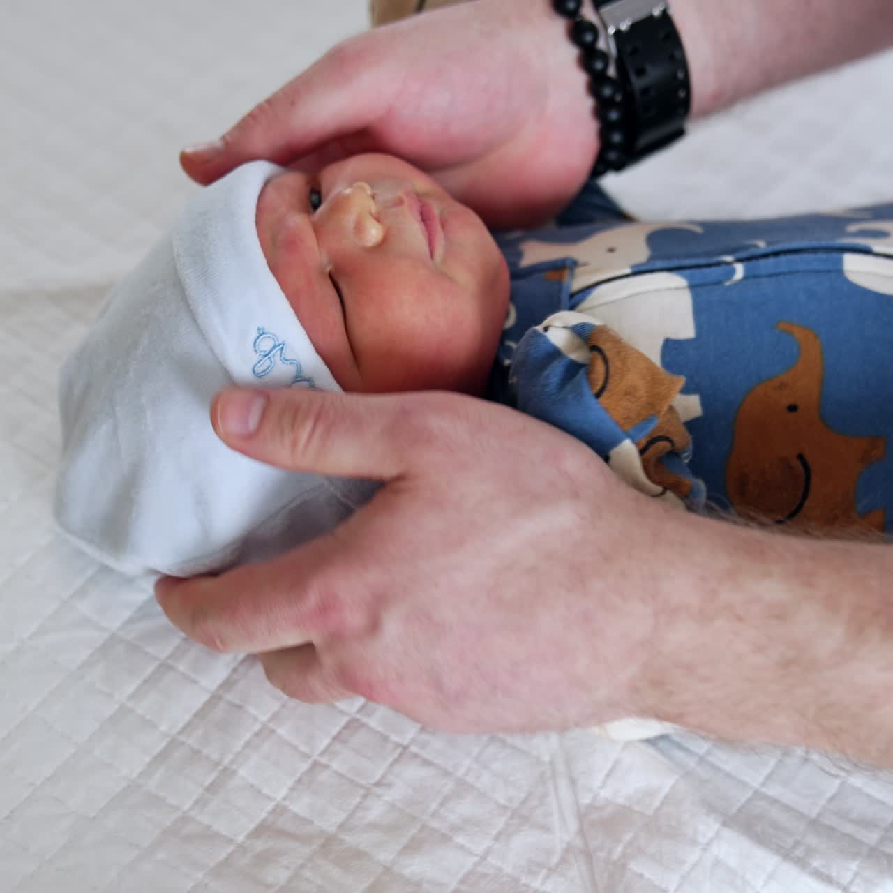 Little Caucasian newborn in a blue romper is put a white cap on. Dad's hands dressing a baby lying on the sheet. Close up