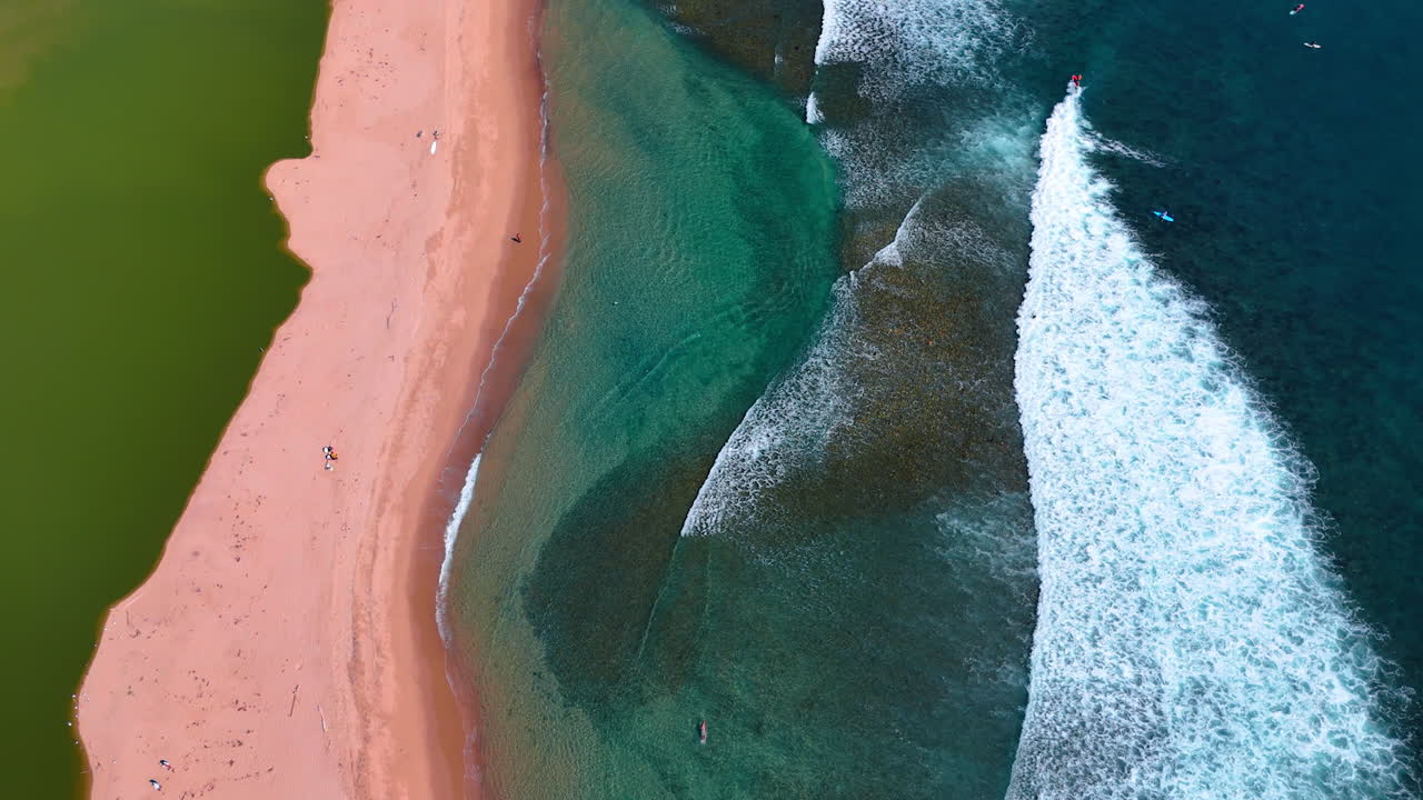 Sea-green water and wide white foamy waves roll to the narrow sandy beach. Few people walk by the shore. Top view