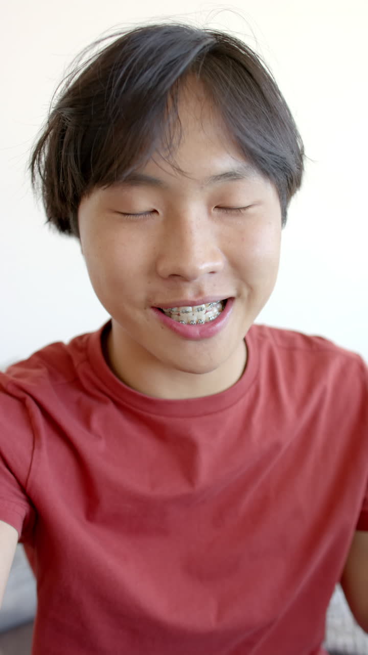 Vertical video: Smiling Asian teenage boy with braces wearing red shirt at home, feeling happy