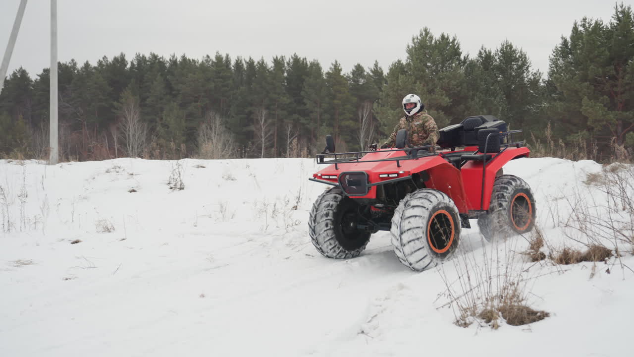 ATV rosso che conquista una collina innevata, pilota mimetizzato con casco si fa strada attraverso un nevaio vicino a una pineta, pneumatici chiodati sovradimensionati che sollevano neve polverosa, primo piano del battistrada del pneumatico, luce fredda e coperta, sport d'azione