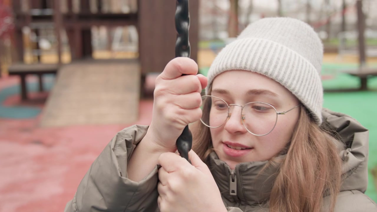 Woman holding swing chain in playground with glasses and knit beanie, adjusting grip and gazing upward, introspective mood with soft winter light and wooden play structure backdrop