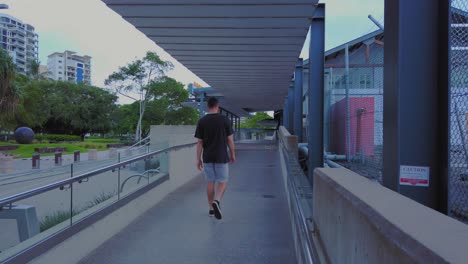 Man-Wearing-Black-Shirt,-Walking-On-The-Street-With-Unique-Buildings-and-Trees---Wide-Shot