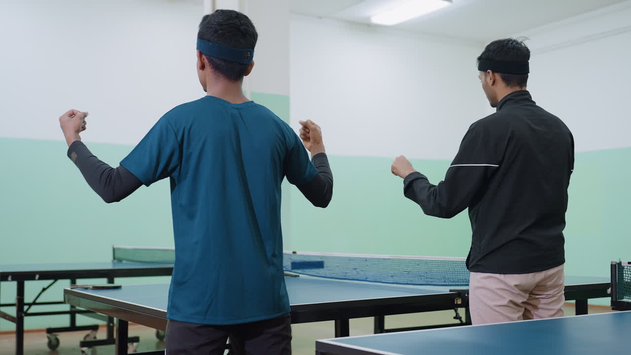 Back view of two friends in sportswear performing wrist flexibility warmup near tennis table, focusing on preparation, coordination, and physical readiness before sports activity inside indoor court
