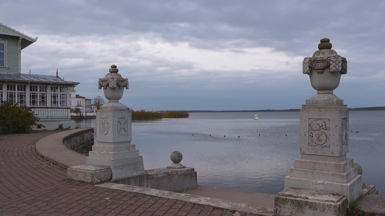 Two engraved stone pillars or statues at seashide by water in Haapsalu Promenade area surrounded by water and red poppy stone road during a cloudy and windy autumn day that is cold. Dark clouds in sky