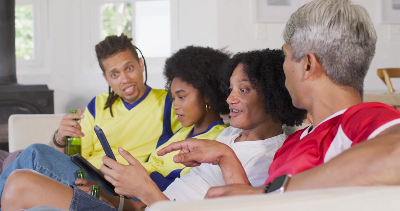 Diverse friends in jerseys leaning in as center man showing phone on couch, watching, holding beers