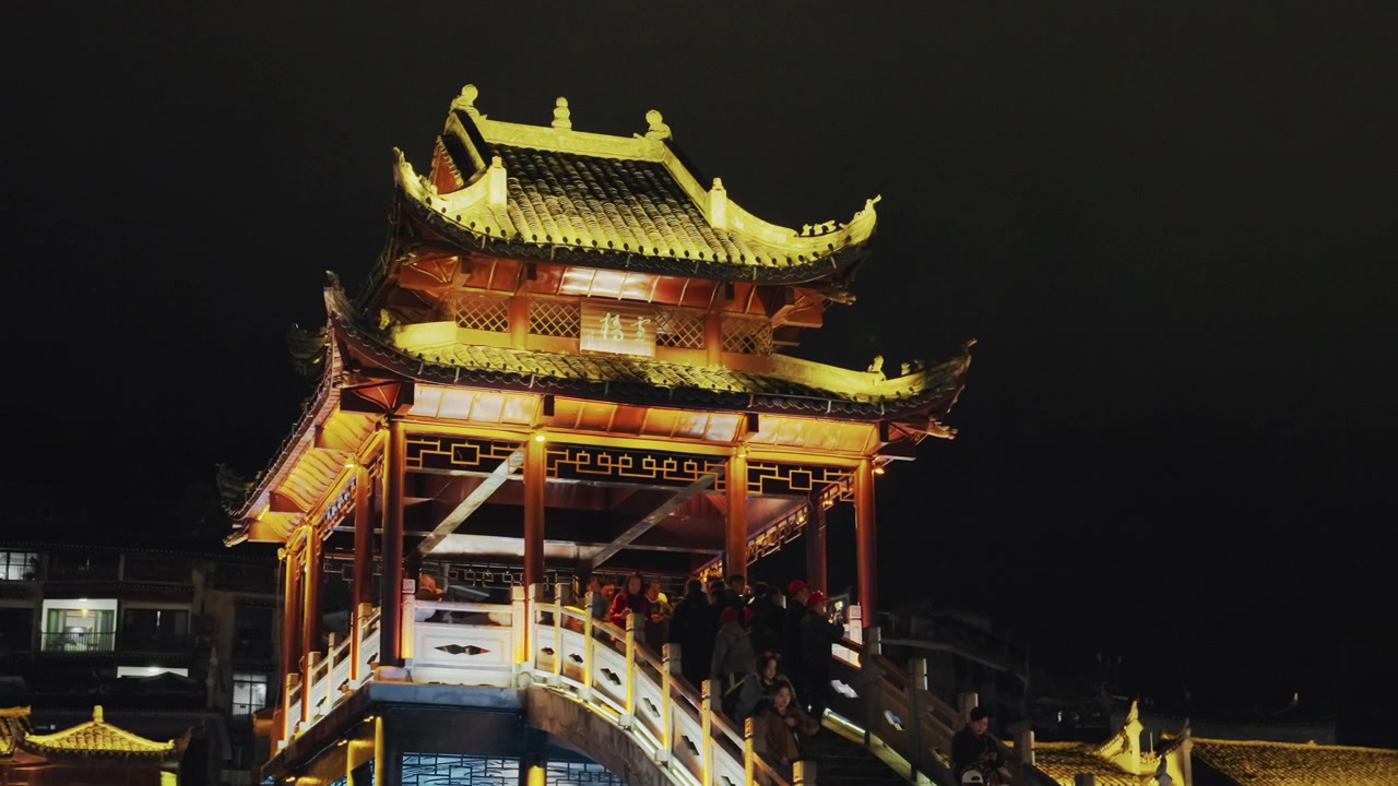 Işıklı pagoda köprüsü gece yürüyüş atmosferi, süslü çatı hatlarını ve kavisli merdivenleri belirleyen sıcak altın fenerler, boş gezinti yolu ve sakin şehir silüeti, gelenekseli çerçeveleyen kasvetli karanlık gökyüzü.
