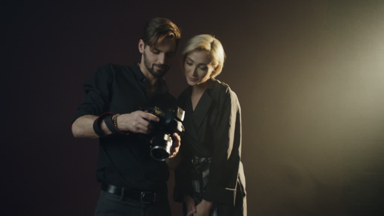 Caucasian male photographer taking a photo of a blond model woman in the dark studio and then shows it to her on the camera screen