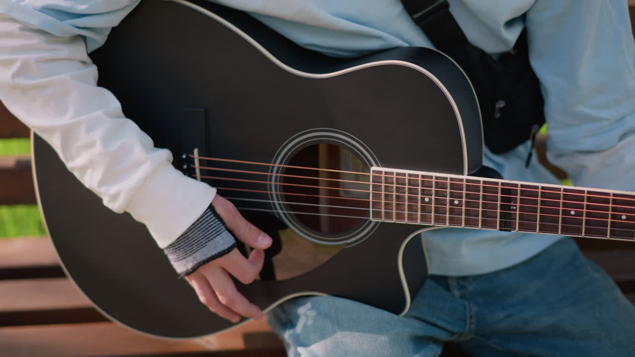 Siedzący gitarzysta grający na czarnej gitarze akustycznej na ławce w parku, zbliżenie dłoni szarpiących struny i tworzących akordy, codzienne dżinsy i lekka kurtka, oświetlone słońcem tło z trawą, łagodny klimat lofi