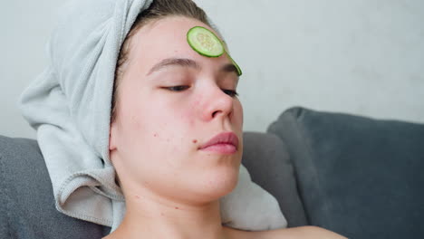 a woman is placing a slice of cucumber on her forehead while relaxing, she has a towel wrapped around her head, and the atmosphere is bright and calm, close-up shot of her facial care routine