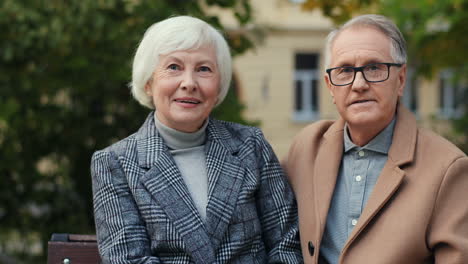 Retrato-De-Una-Pareja-Mayor-Sentada-En-El-Banco-Sonriendo-A-La-Cámara-En-El-Parque-En-Otoño