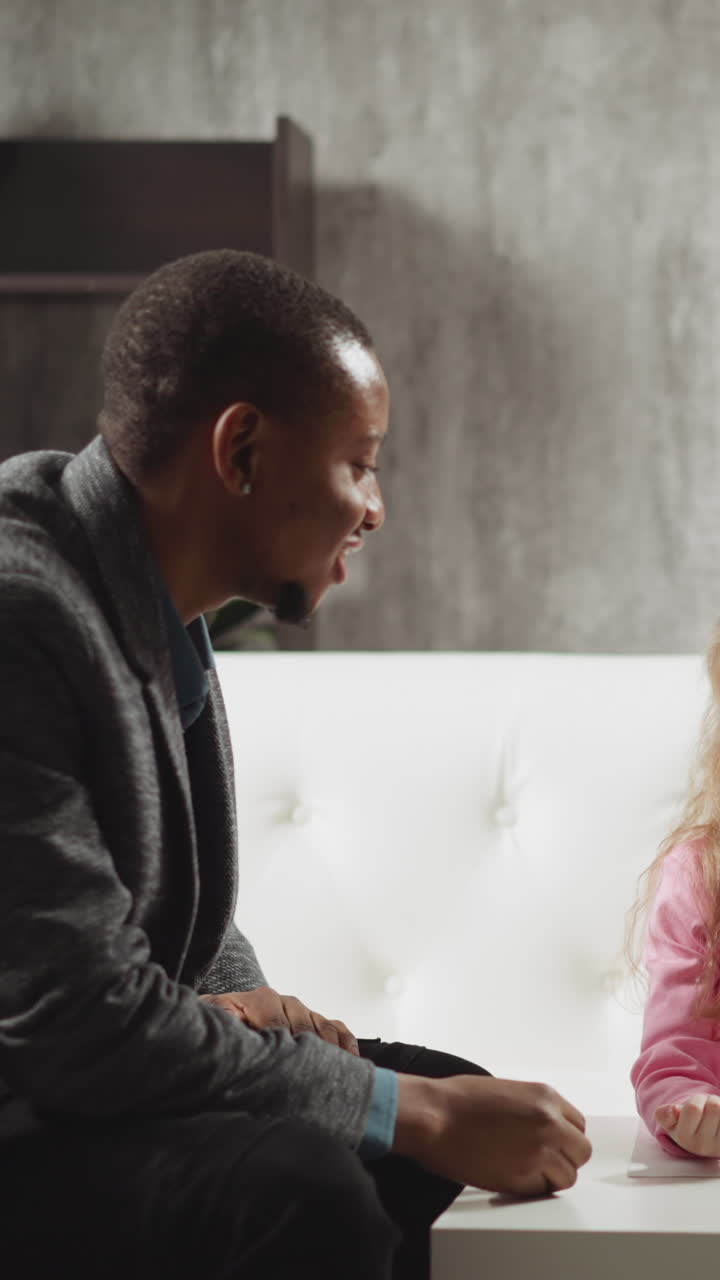 Little girl pupil answers nodding African-American tutor while classmate sits on sofa at home English lesson with native speaker slow motion