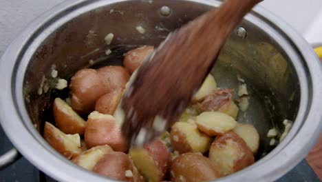 cooking and stirring new potatoes with a large wooden spoon simmering in garlic butter in a stainless steel saucepan, close up of delicious potatoes with red skin on